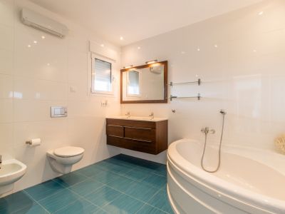 Salle de bain lumineuse avec baignoire d'angle, double vasque en bois foncé et WC suspendu. Carrelage blanc contrastant avec un sol turquoise vif, lumière naturelle et climatisation.