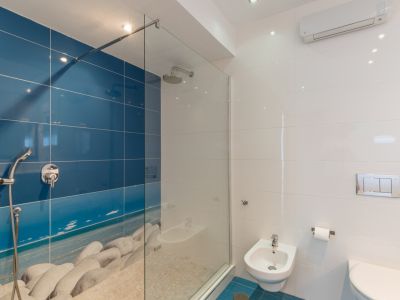 Salle de bain moderne avec douche à l'italienne et paroi vitrée, mur carrelé bleu avec motif plage de galets et sol en pierre. Comprend bidet et WC suspendu.