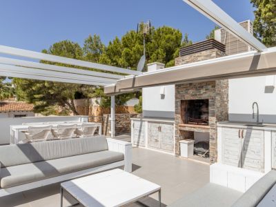 Solarium sur le toit avec cuisine d'été en pierre, barbecue intégré et évier, ainsi que des espaces détente et repas sous une pergola blanche, entouré d'arbres luxuriants.