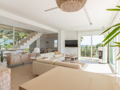 Vaste salon ouvert avec canapé crème, poêle à granulés et balcon avec vue. Une élégante cage d'escalier en verre et un meuble en bois rustique sont visibles.