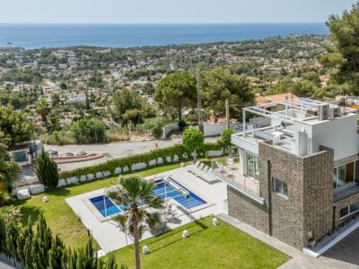 Vue aérienne de la villa moderne avec revêtement en pierre, piscine rectangulaire, pelouse soignée et solarium sur le toit avec pergola, offrant des vues mer panoramiques.