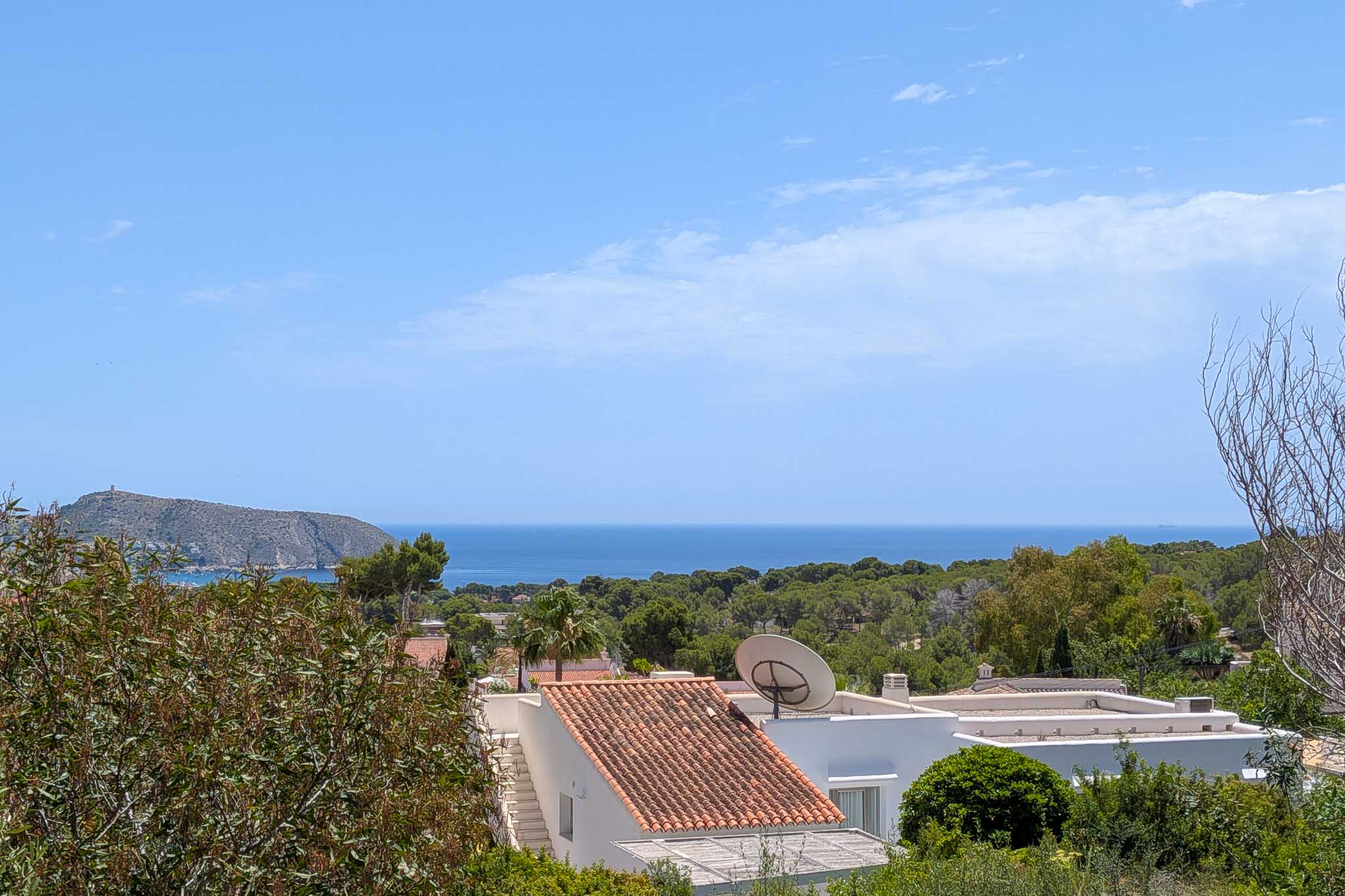Vue élevée sur la Méditerranée, avec des toits en tuiles traditionnelles et une végétation luxuriante au premier plan. Un promontoire rocheux avec une tour lointaine sous un ciel bleu clair.