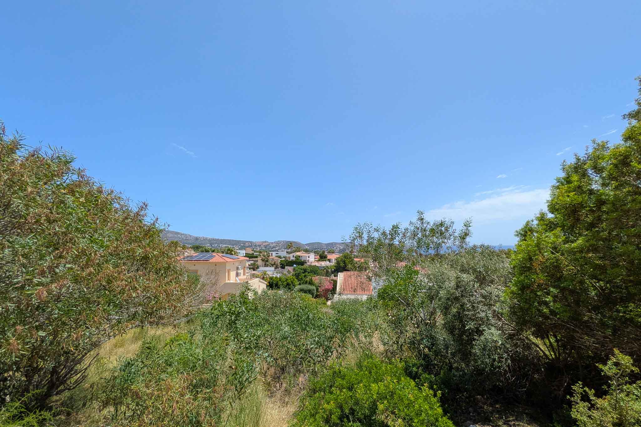 Vue panoramique depuis la parcelle, dévoilant un quartier résidentiel verdoyant avec toits traditionnels, la mer lointaine et les montagnes sous un ciel bleu clair.