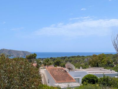 Vue élevée sur la Méditerranée, avec des toits en tuiles traditionnelles et une végétation luxuriante au premier plan. Un promontoire rocheux avec une tour lointaine sous un ciel bleu clair.