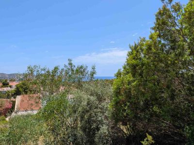 Vue panoramique depuis la parcelle, avec une végétation méditerranéenne luxuriante au premier plan, des toits résidentiels et la mer Méditerranée à l'horizon sous un ciel clair.