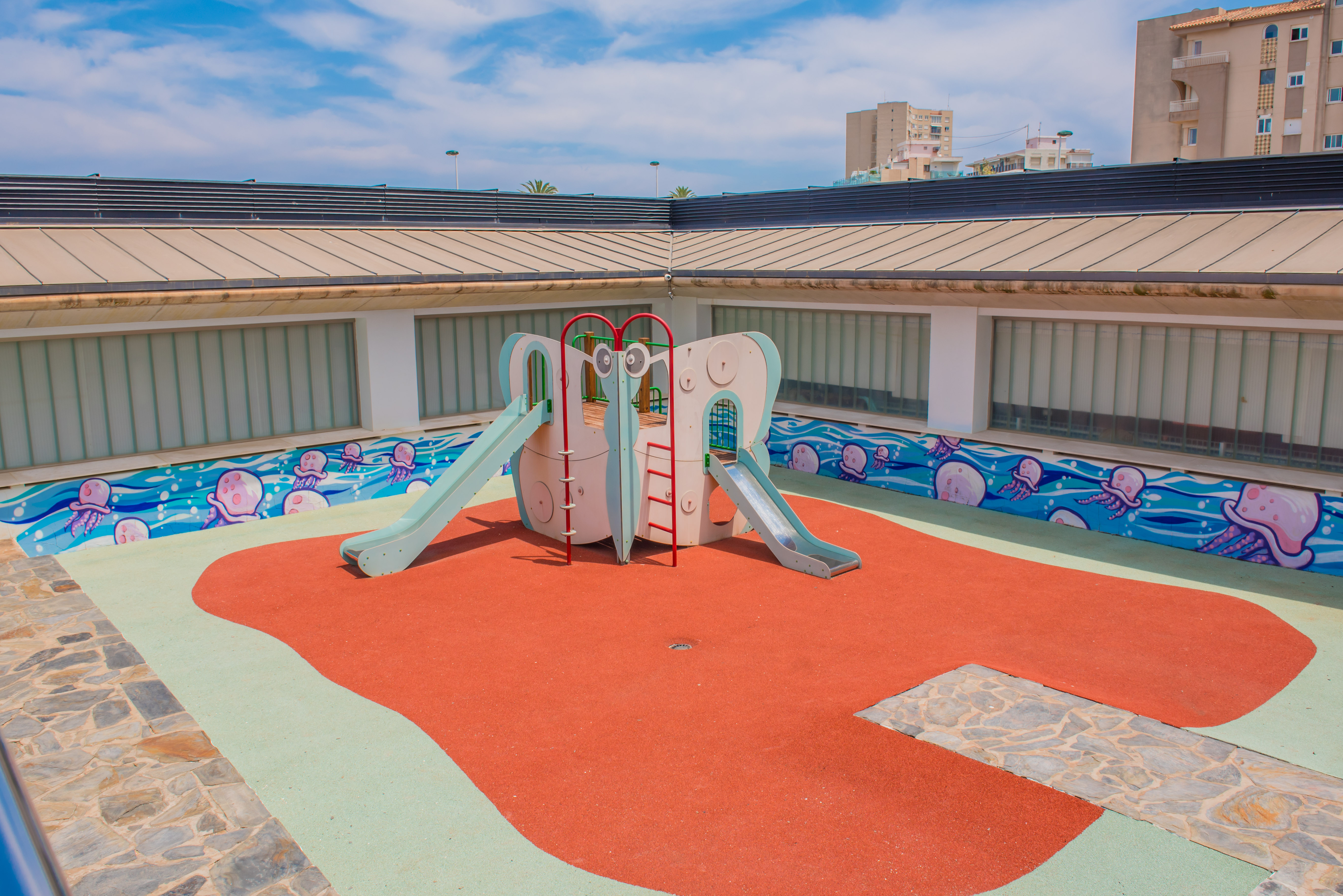 Aire de jeux pour enfants lumineuse avec structure de toboggans sur un sol en caoutchouc rouge et vert. Un mur décoré d'une fresque de méduses anime l'espace, sous des panneaux de verre dépoli.