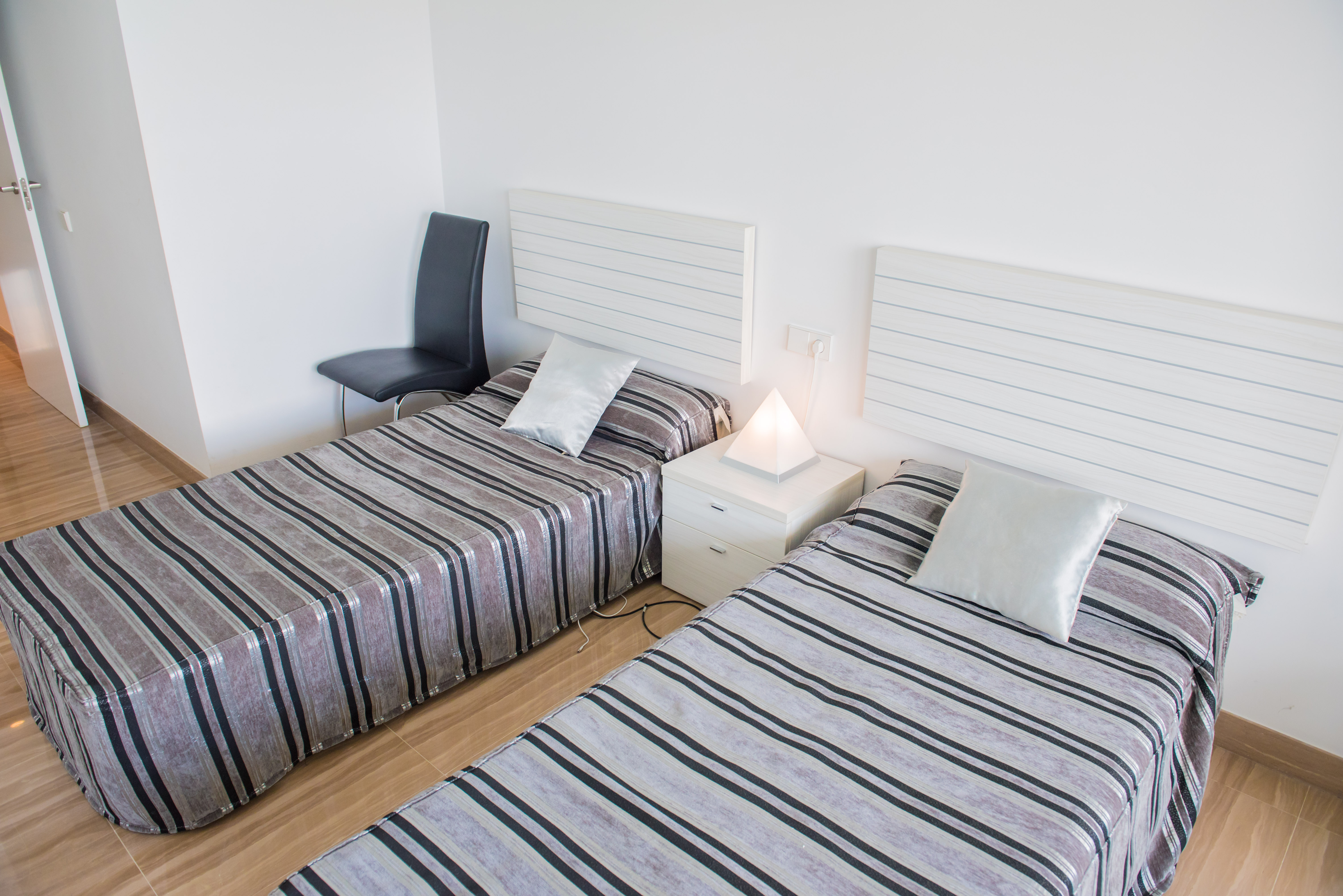 Chambre avec deux lits simples, têtes de lit blanches à lattes et literie rayée. Une lampe pyramidale éclaire la table de chevet située entre les lits.