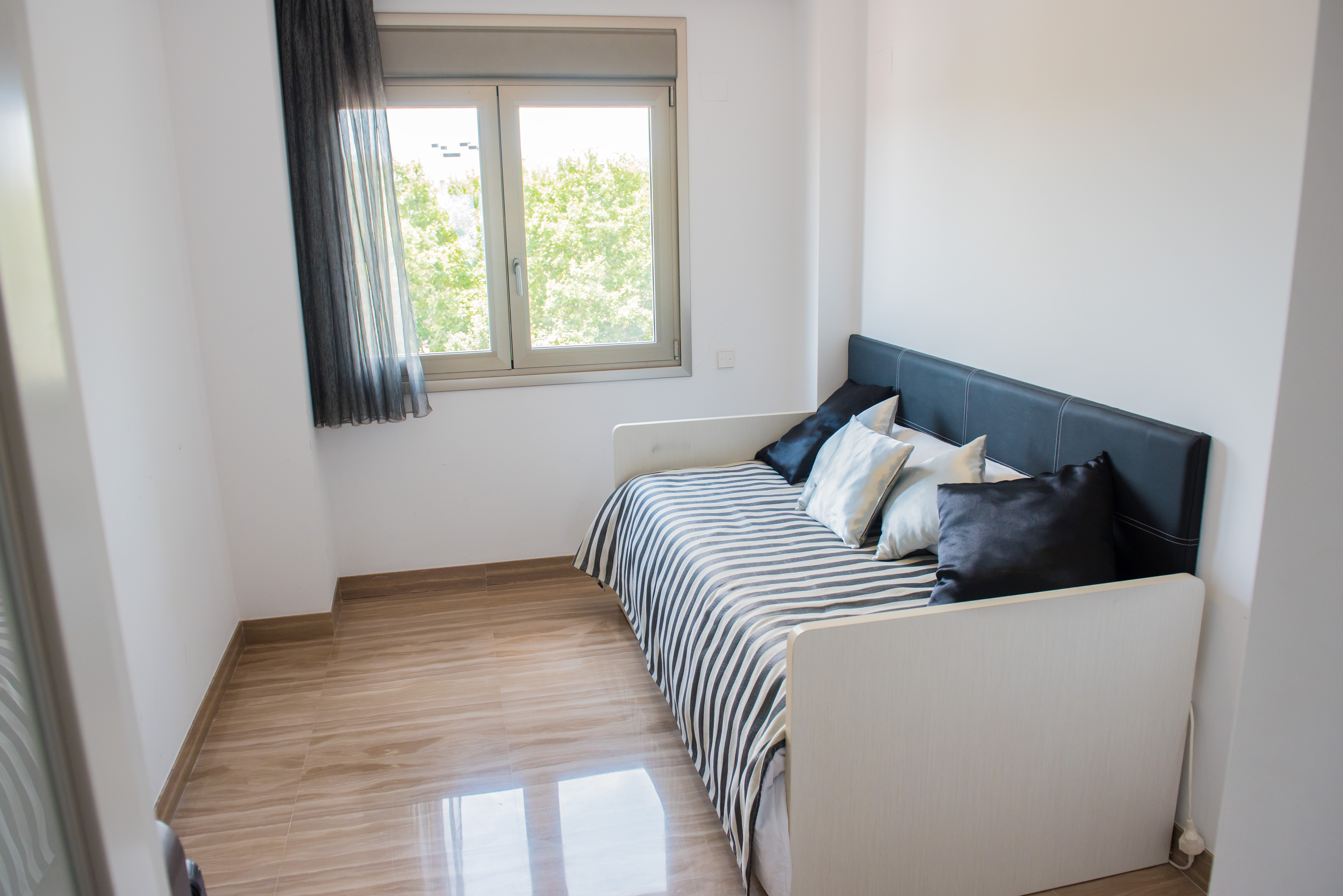 Chambre individuelle avec lit de jour, tête de lit noire capitonnée et literie rayée. Le sol en marbre clair poli et la fenêtre offrent une vue sur la verdure.