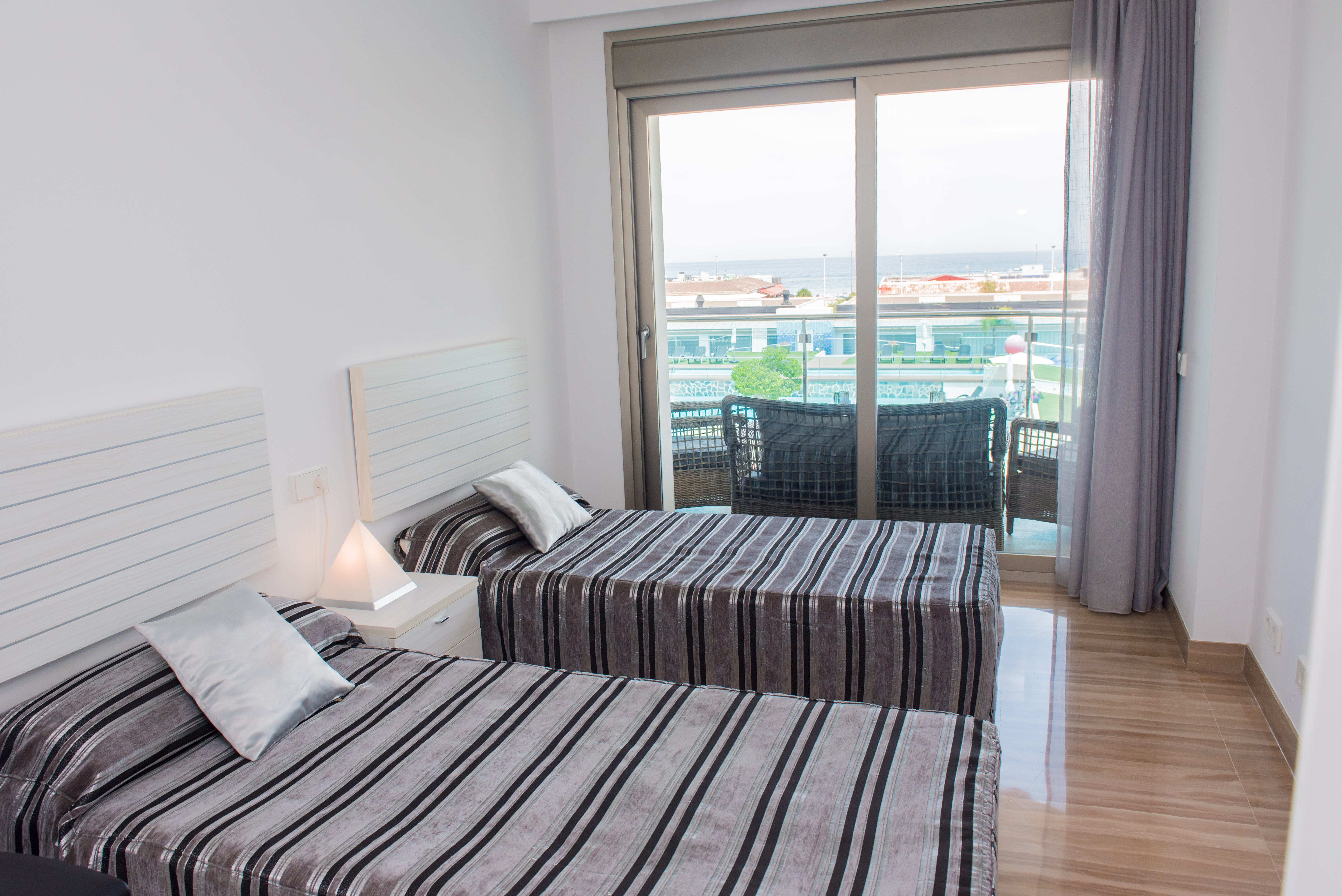 Chambre lumineuse avec deux lits simples, literie rayée et table de chevet. Des portes coulissantes mènent à un balcon avec sièges extérieurs et vue partielle sur la mer. Sol poli.