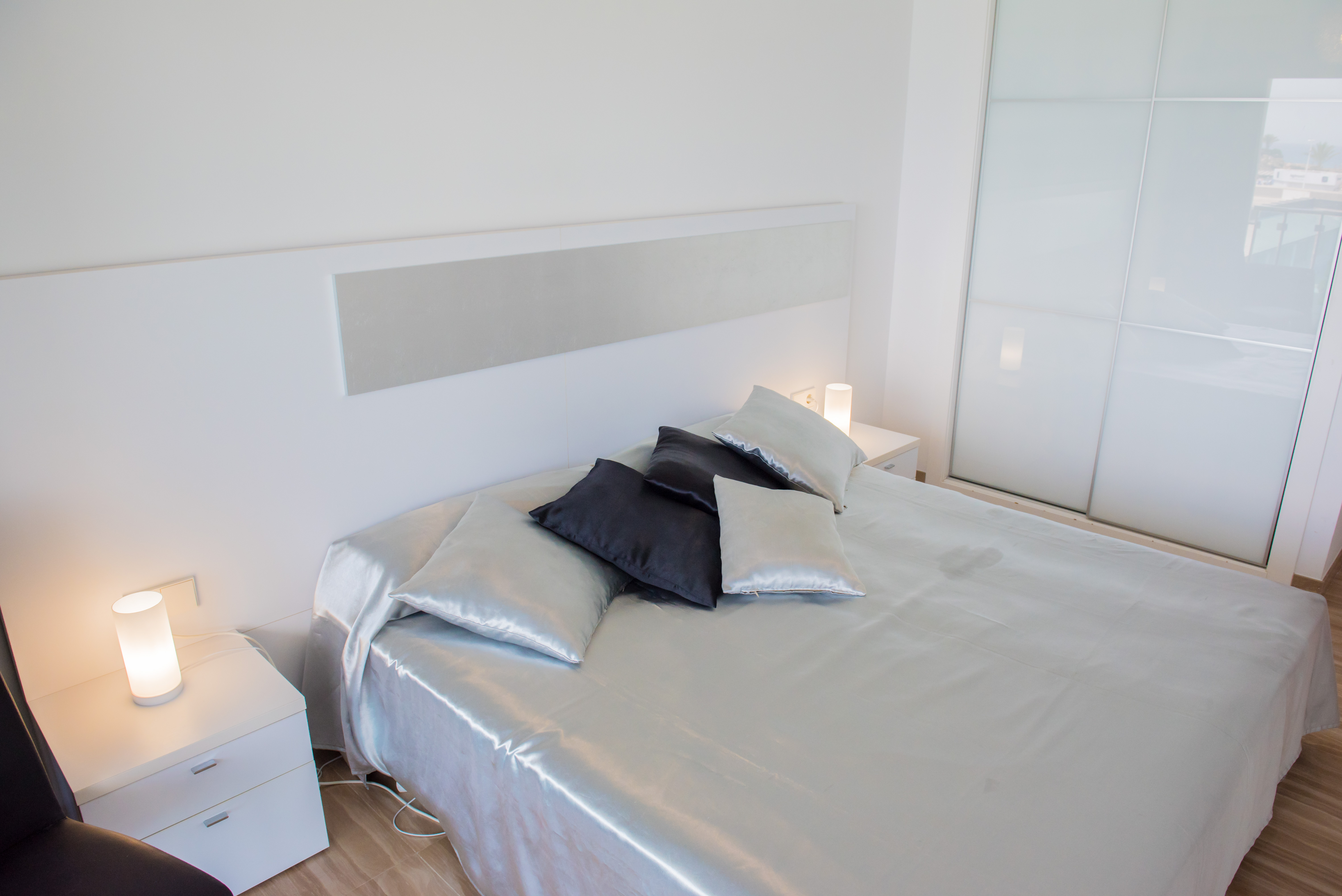 Chambre moderne avec lit aux draps satinés argentés, tête de lit blanche et tables de chevet assorties avec lampes cylindriques. Armoire coulissante en verre dépoli.