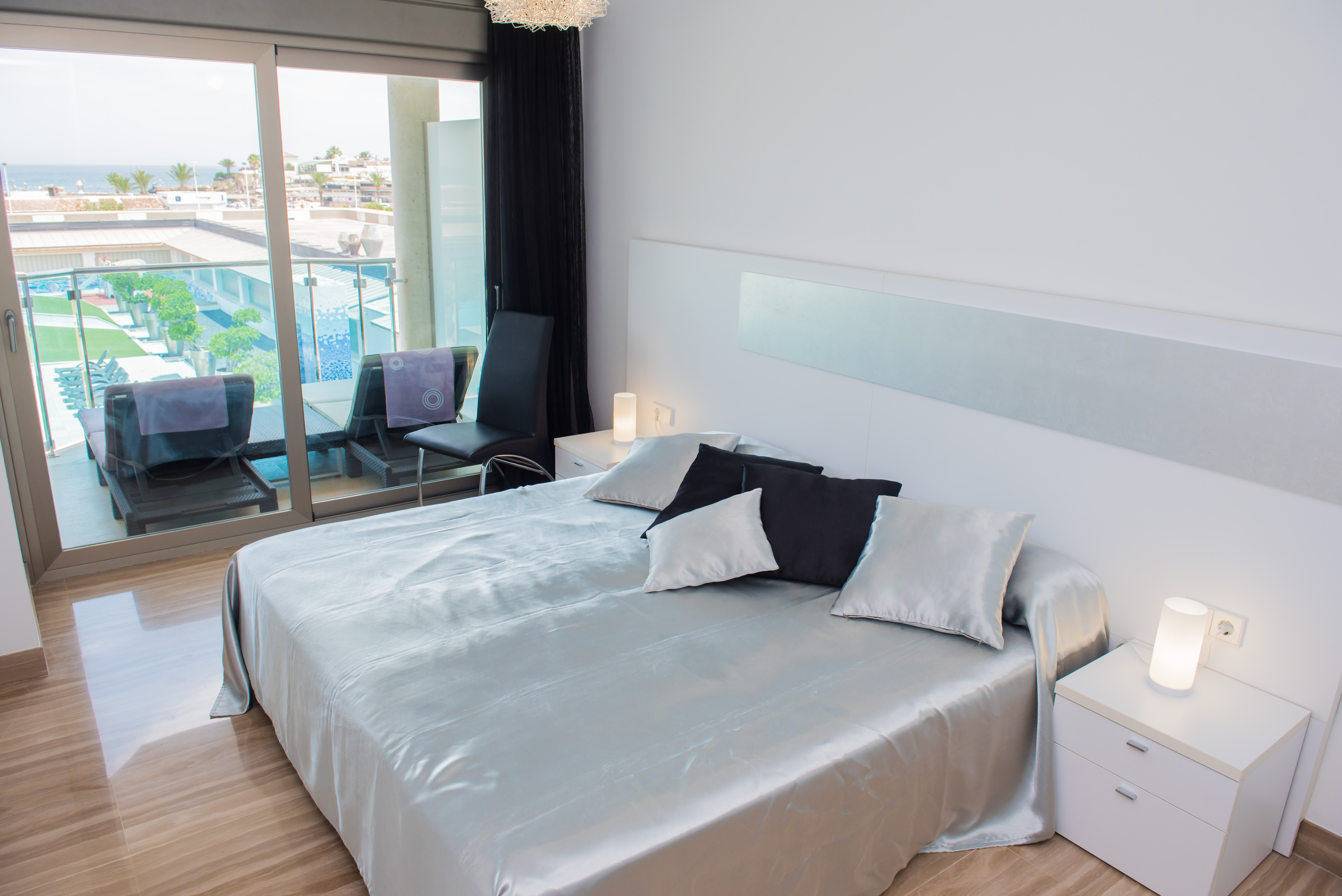 Chambre principale moderne avec grand lit, literie satinée argentée, carrelage brillant et portes coulissantes vers la terrasse offrant une vue sur la mer et la piscine.