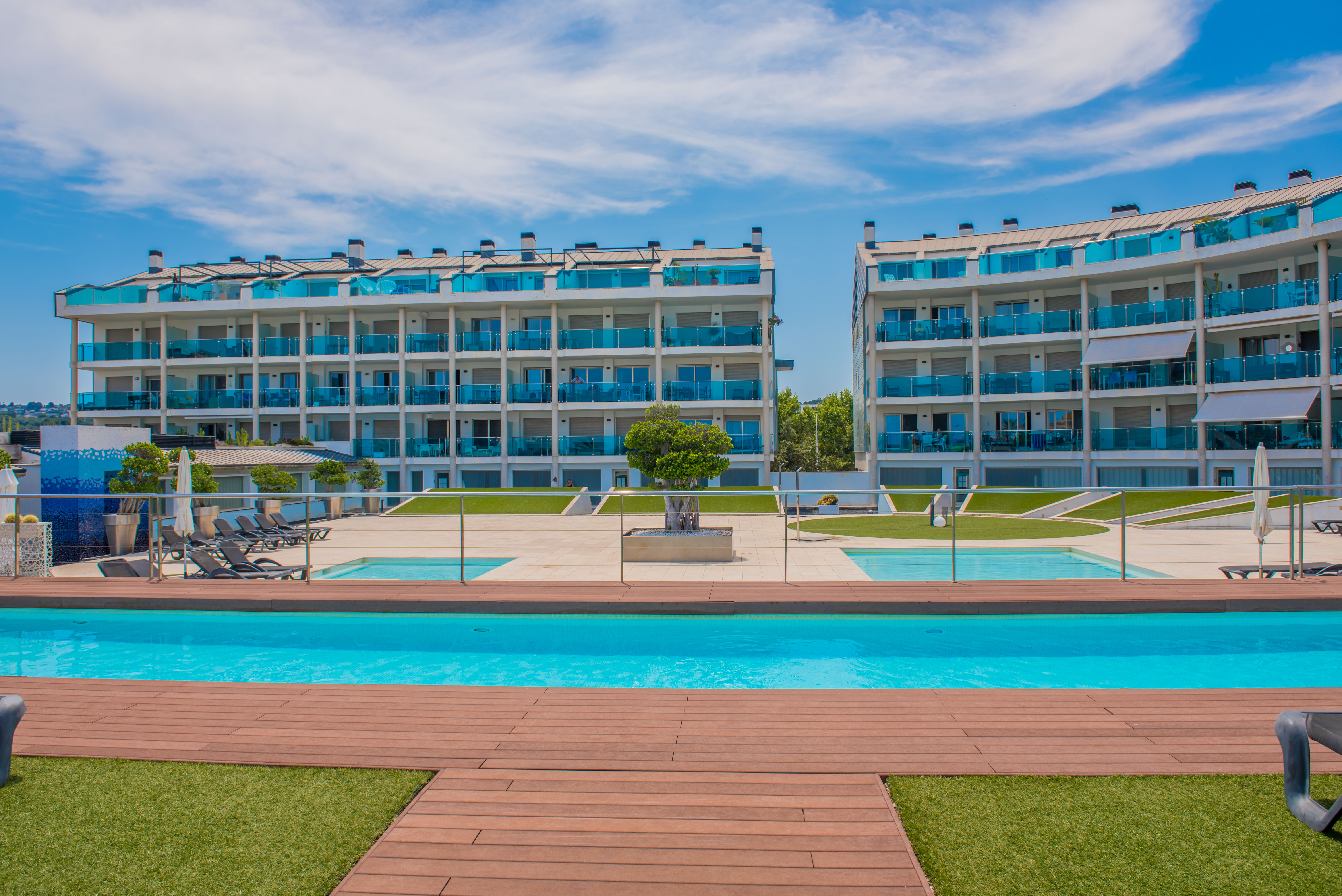 Complexe résidentiel moderne composé de deux bâtiments avec balcons incurvés en verre bleu, entourant une grande piscine commune avec terrasse en bois élégante et gazon artificiel.