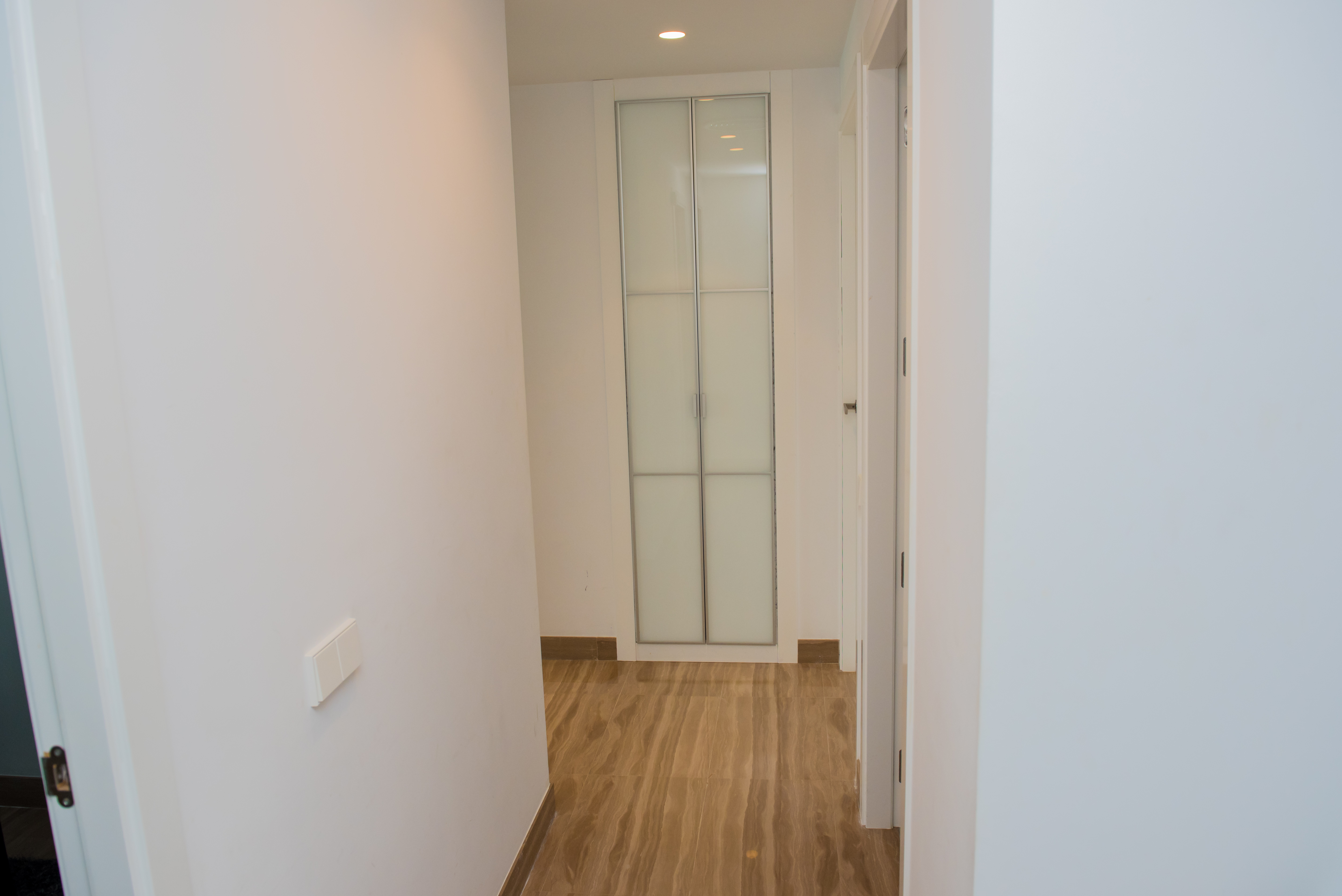 Couloir intérieur aux murs blancs et sol carrelé effet marbre veiné. Au fond, une armoire encastrée avec portes en verre dépoli et cadre argenté, éclairage encastré.