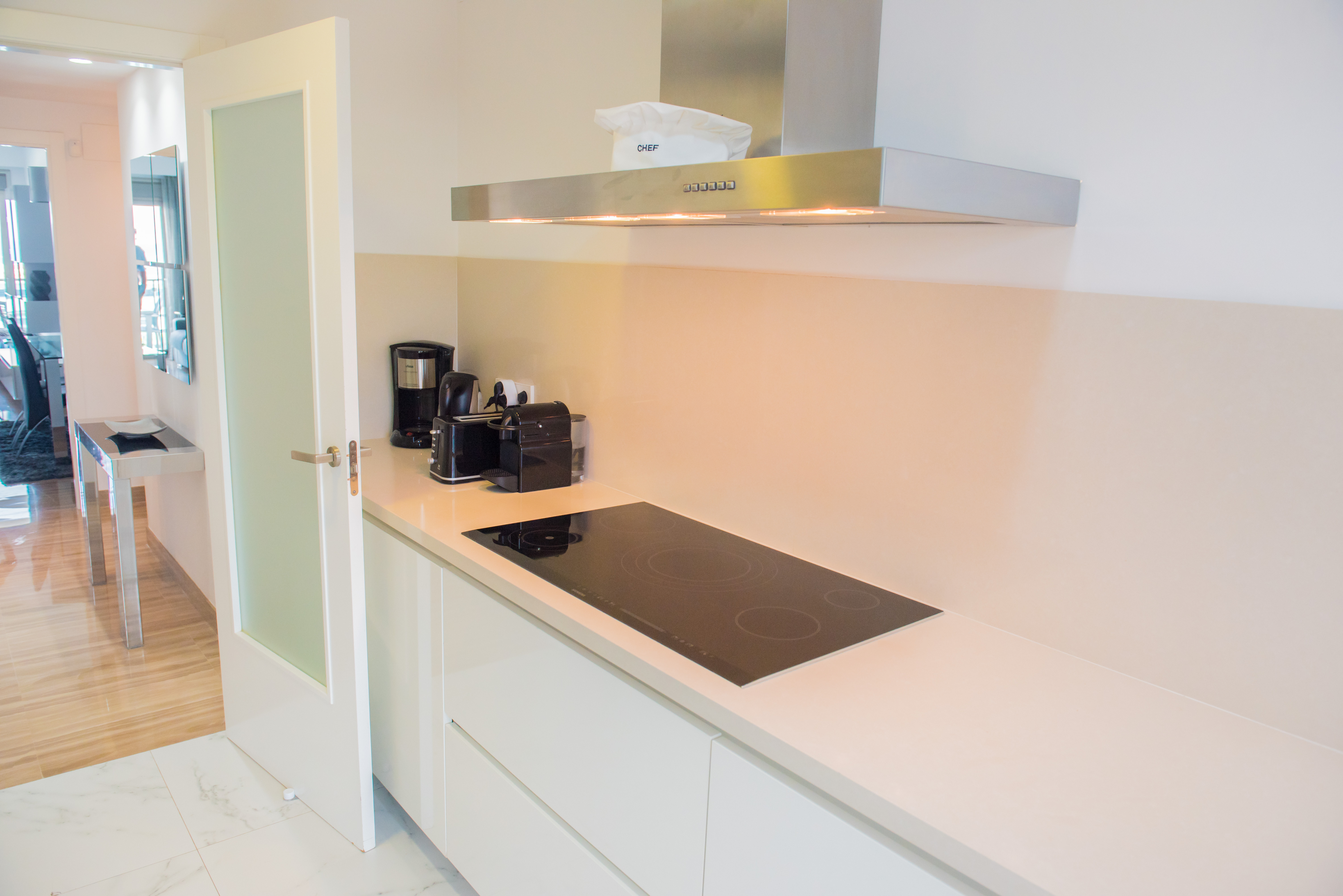 Cuisine moderne avec plan de travail clair, plaque à induction noire et hotte aspirante en acier inoxydable. Le sol carrelé effet marbre s'ouvre sur un espace avec parquet.