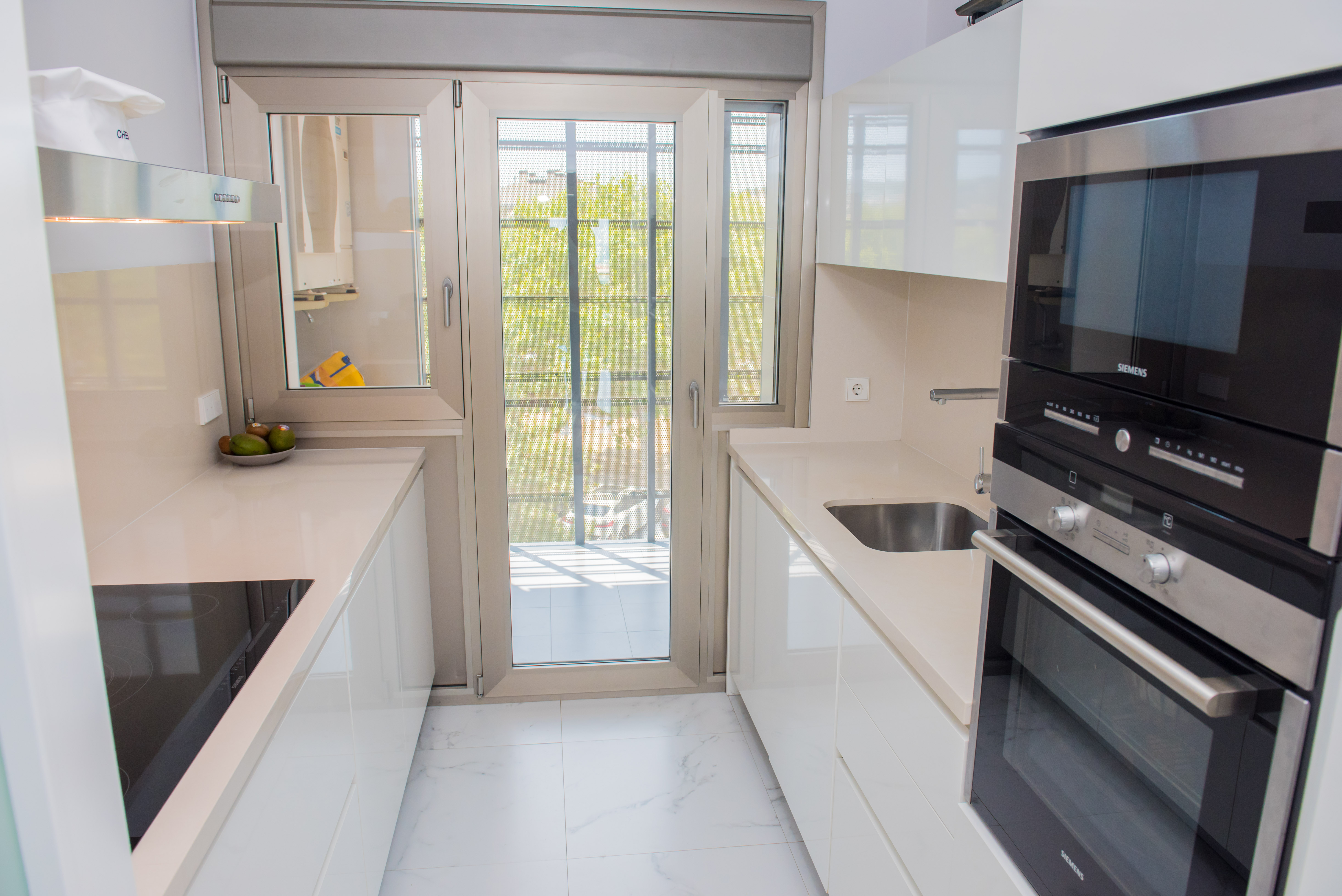 Cuisine moderne en couloir avec des armoires blanches laquées, des plans de travail clairs et des appareils Siemens intégrés. Une porte vitrée double battant offre lumière naturelle et accès au balcon.