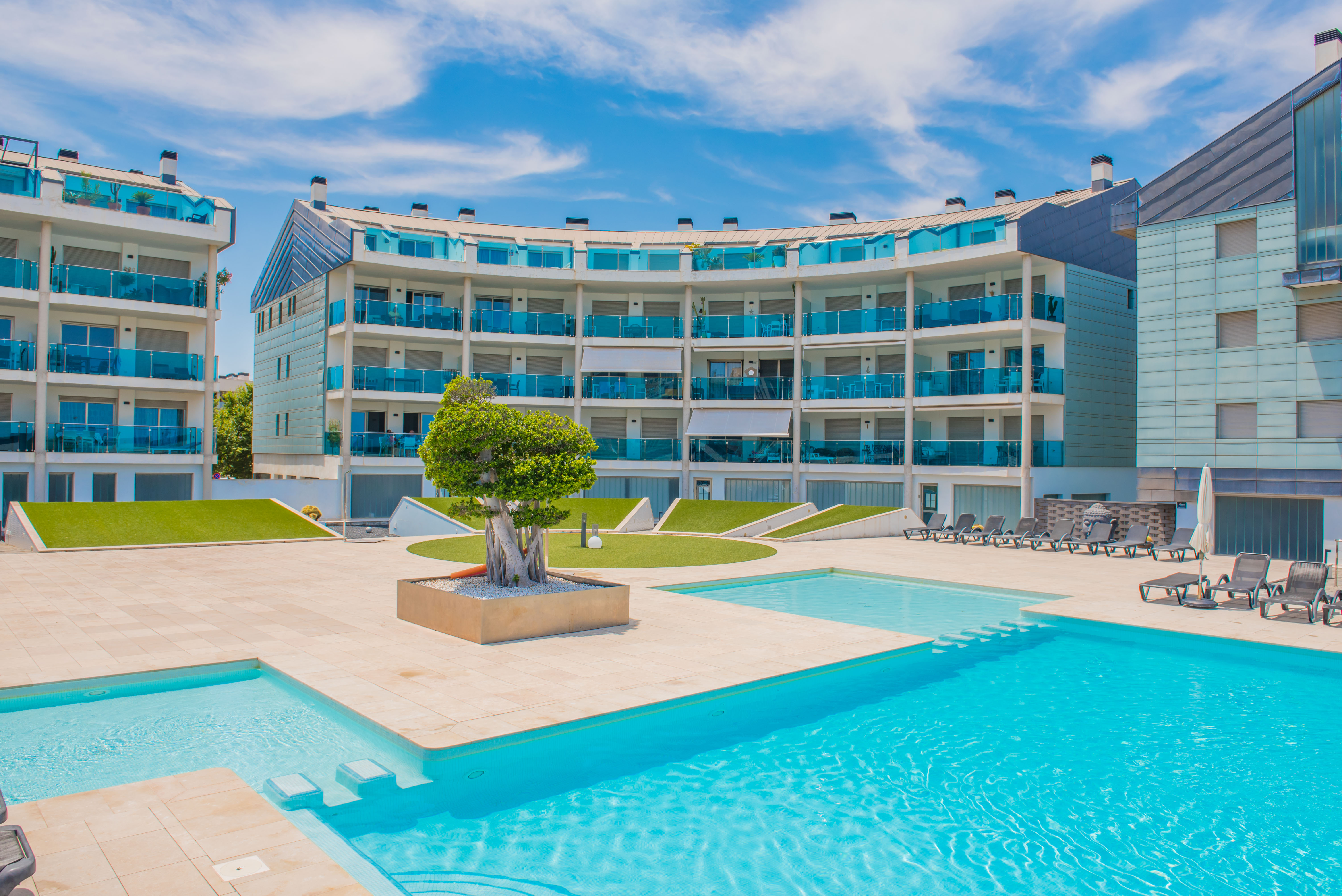 Espace piscine communautaire avec dallage clair, chaises longues et bâtiments résidentiels modernes aux balcons courbés en verre bleu.