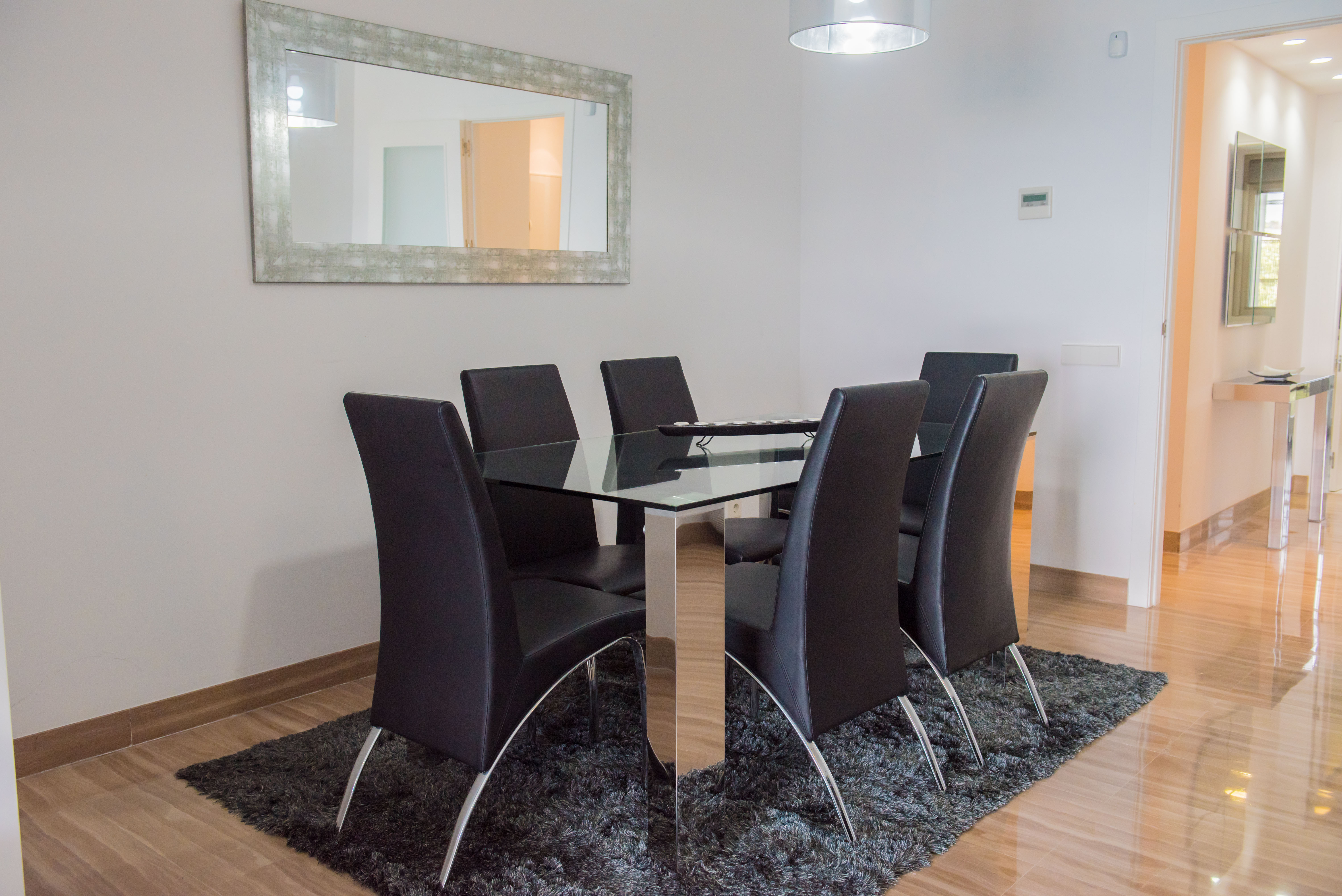 Espace repas contemporain avec table en verre et six chaises en similicuir noir. Un miroir vieilli et un tapis gris complètent le sol en marbre poli.