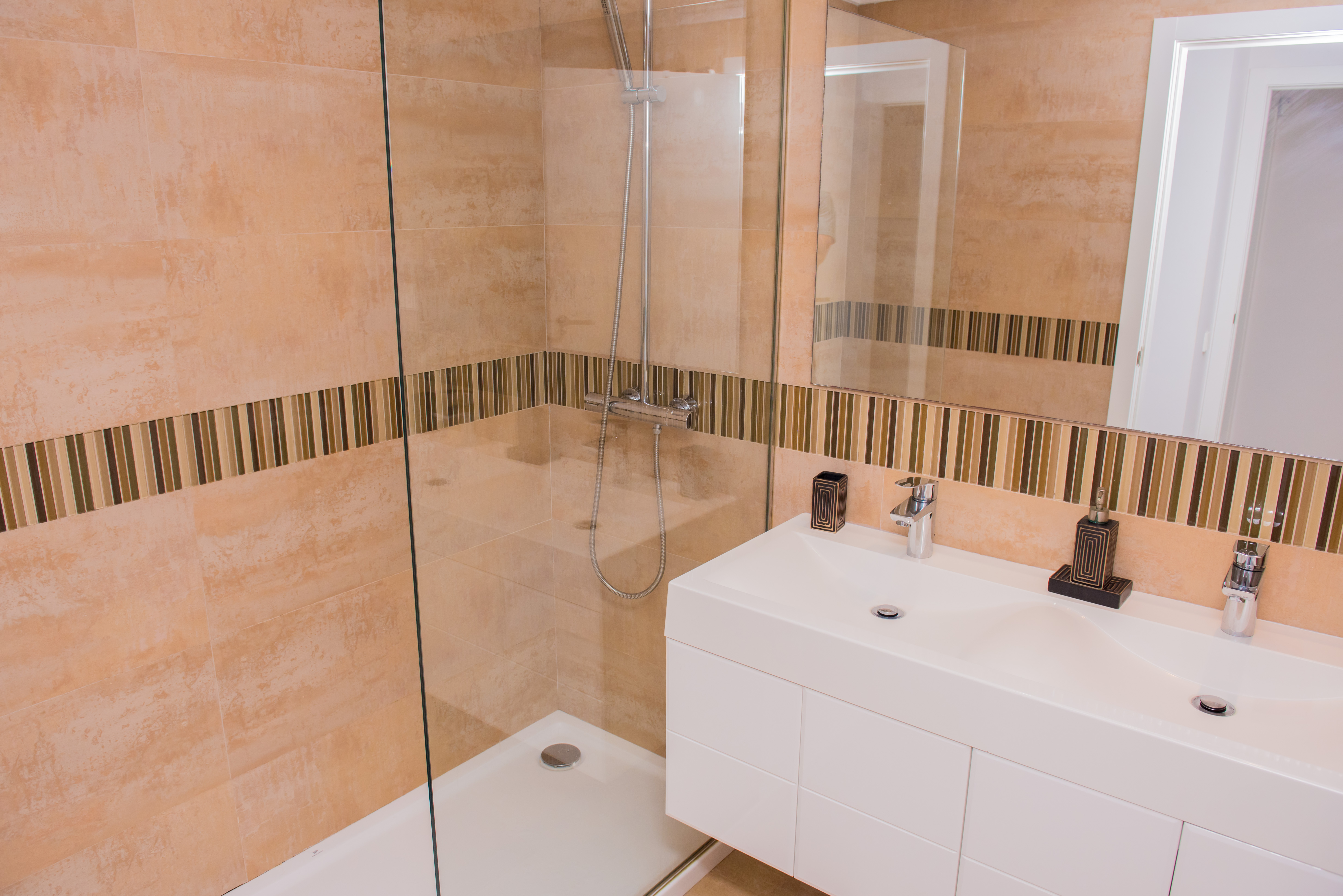 Salle de bain moderne avec douche à l'italienne vitrée et double vasque. Les carreaux muraux beiges sont rehaussés d'une bande décorative en mosaïque.
