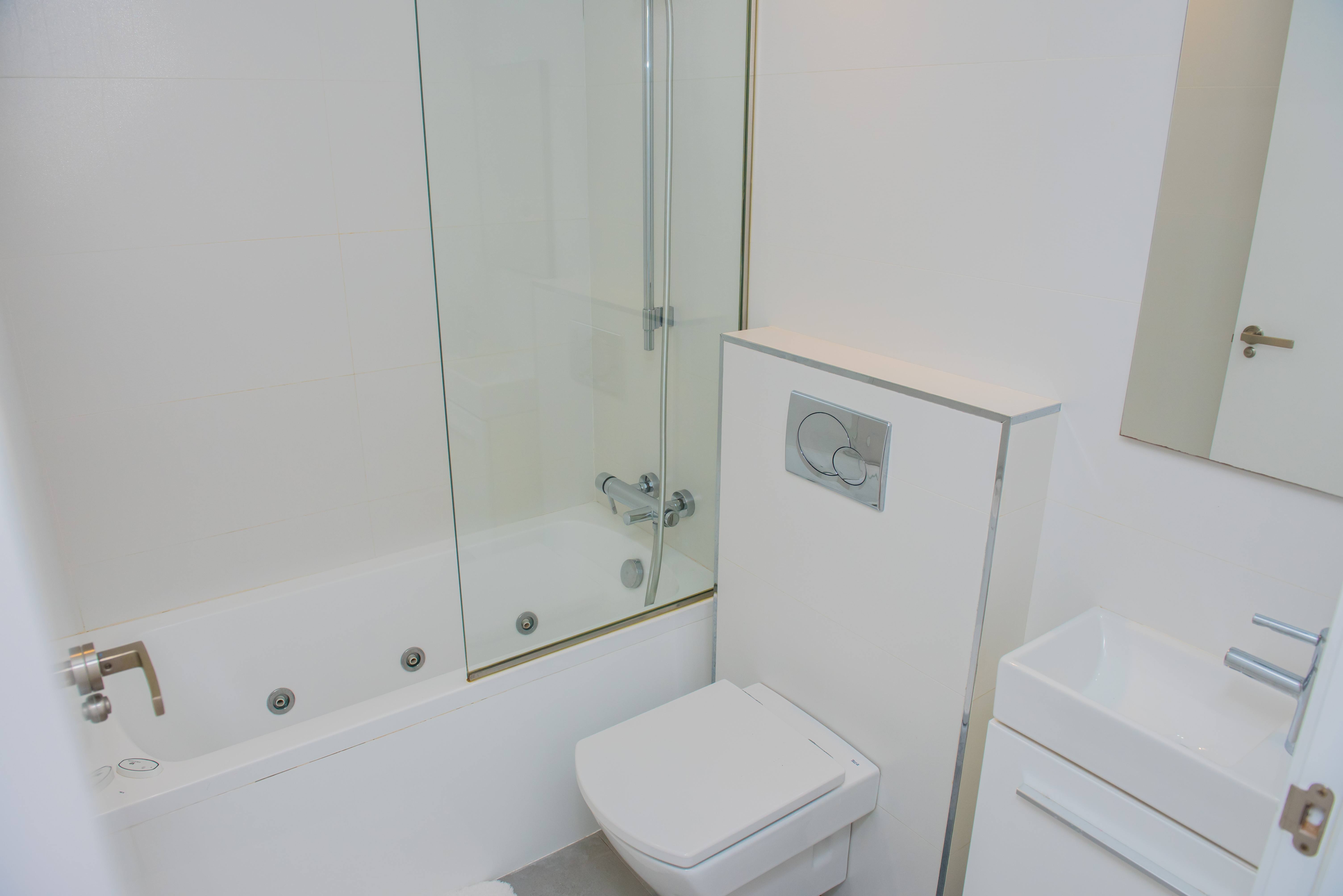 Salle de bain moderne blanche avec baignoire à jets intégrés, paroi de douche en verre, WC suspendu et lavabo rectangulaire. Carrelage mural blanc et robinetterie chromée.