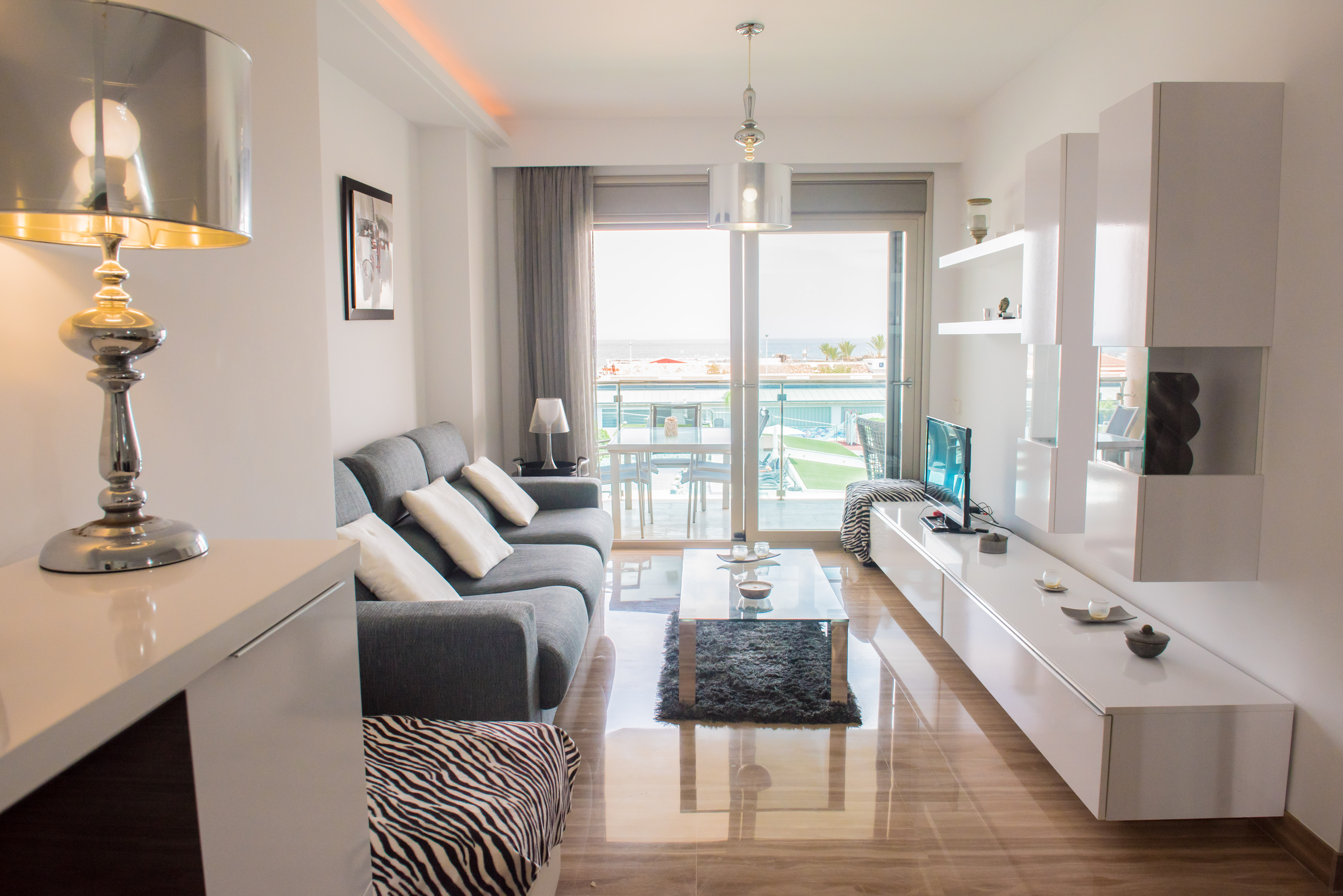 Salon moderne avec sols en marbre poli, s'ouvrant sur une terrasse offrant des vues sur la piscine commune et la mer. Canapé gris et meubles blancs contemporains.