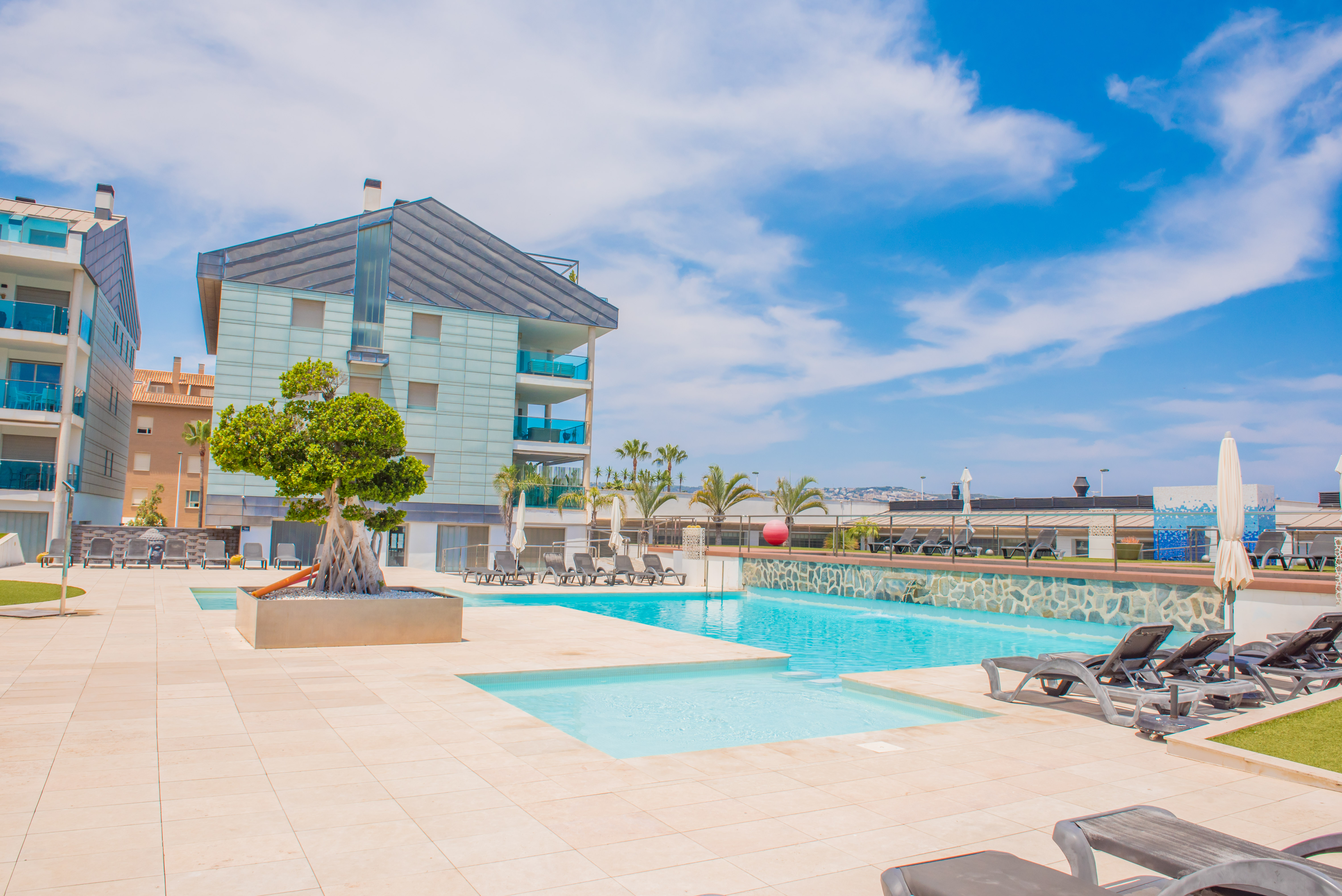Vaste espace piscine commun avec deux bassins distincts, pavage clair et nombreuses chaises longues. Des bâtiments résidentiels modernes aux balcons vitrés bleus surplombent cette zone ensoleillée.
