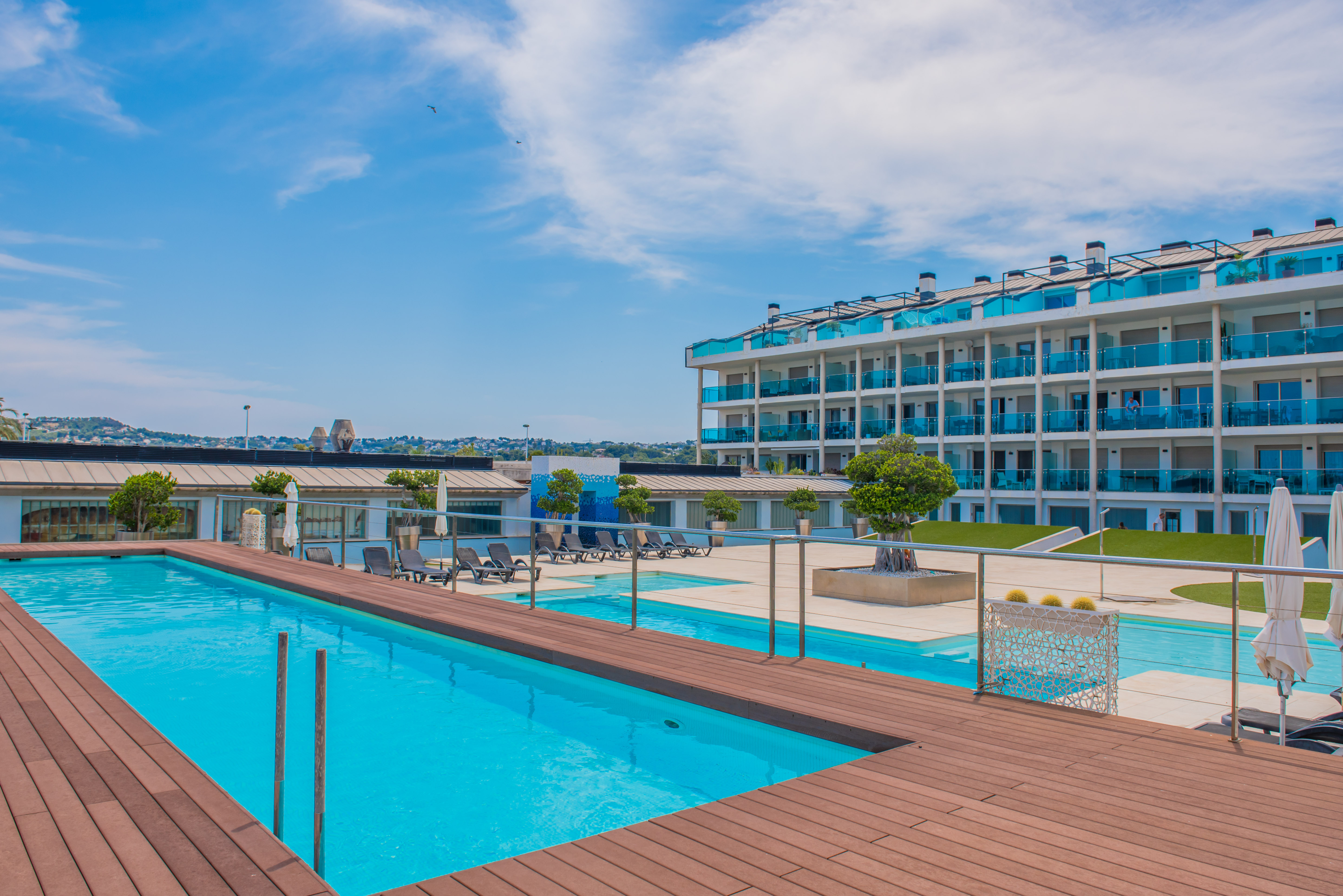 Vaste espace piscine commun avec une piscine d'un bleu éclatant bordée d'une terrasse en bois chaleureuse. L'immeuble moderne aux balcons en verre bleu distinctifs surplombe cet espace extérieur serein.