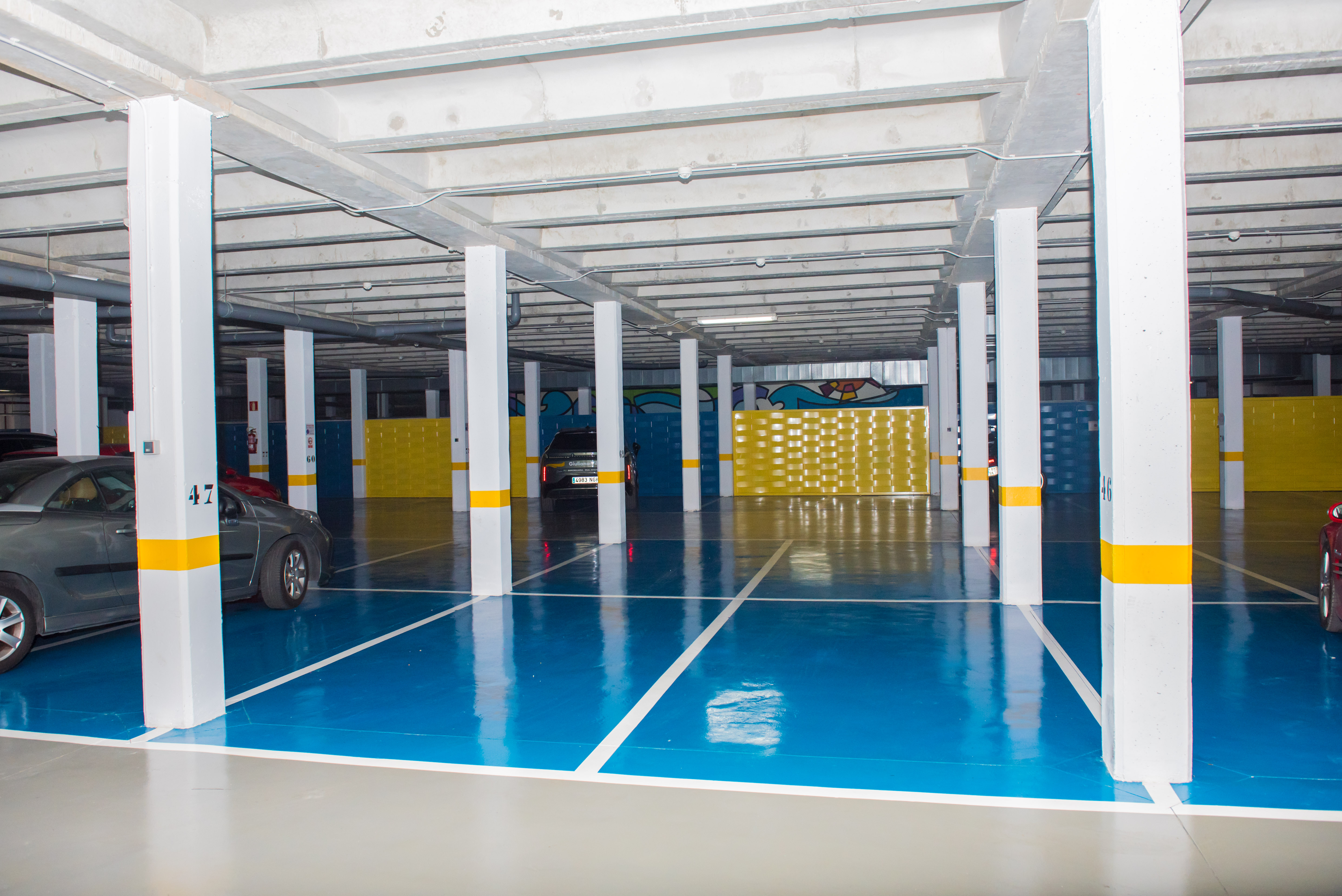 Vaste garage souterrain avec sol en résine bleu et gris, piliers blancs à bandes jaunes et plafond en béton apparent. Des places de stationnement et des panneaux décoratifs jaunes sont visibles.