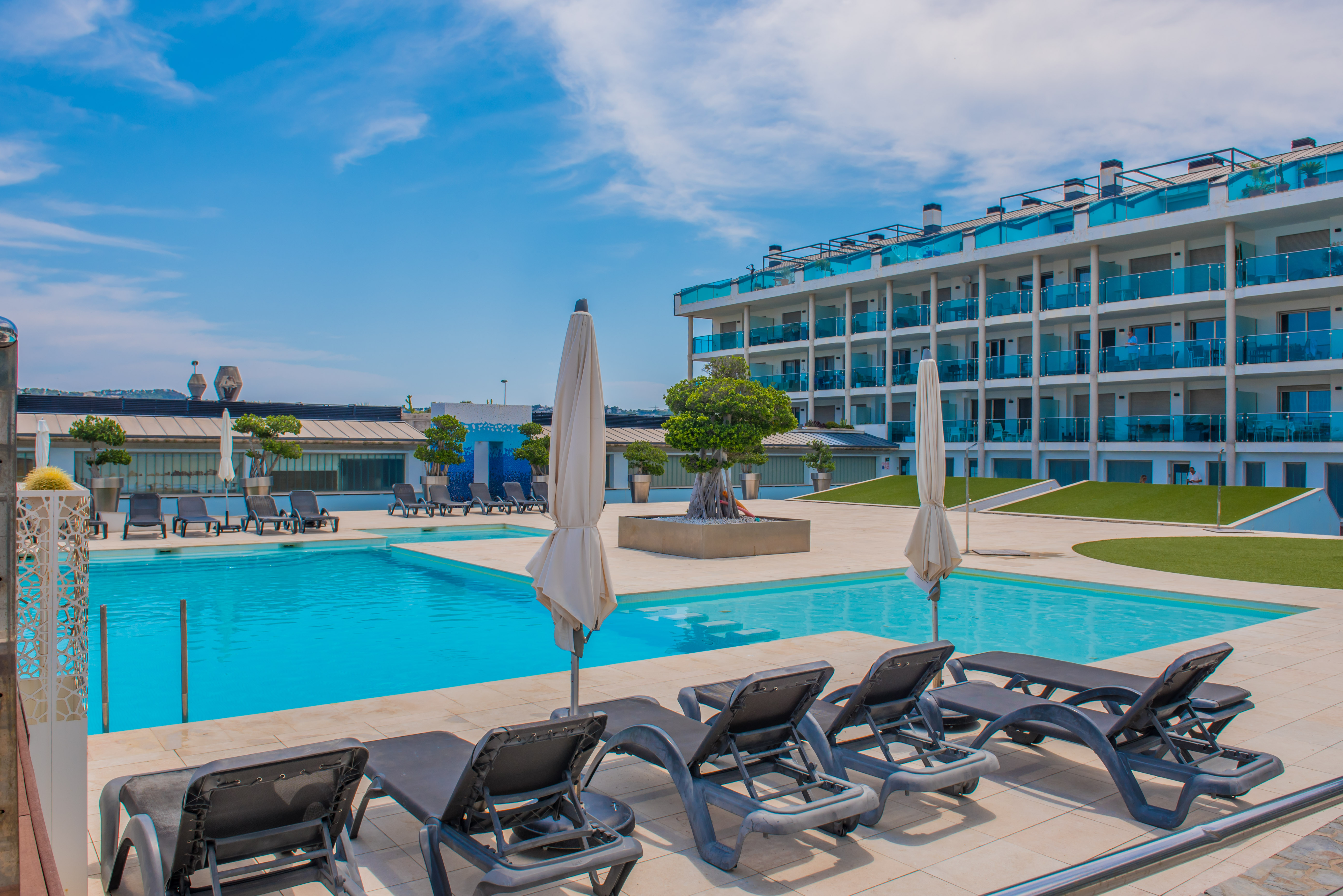 Vaste piscine commune avec chaises longues sur une terrasse carrelée, bordée par un immeuble moderne aux balcons équipés de garde-corps en verre bleu.