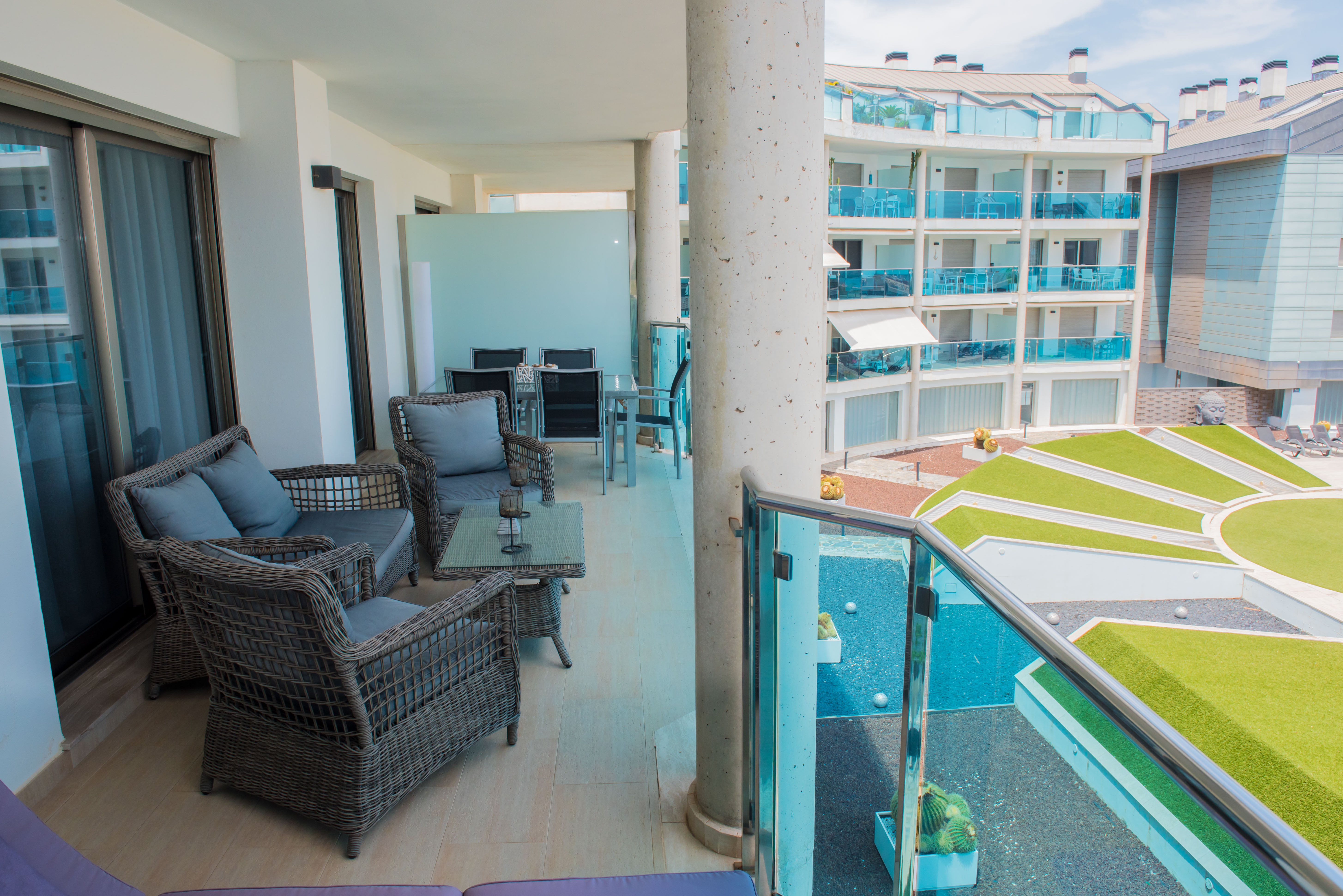 Vaste terrasse couverte avec salon en rotin, table basse en verre et carrelage clair. Vue sur les jardins communs modernes et un bâtiment voisin aux balcons en verre bleu.