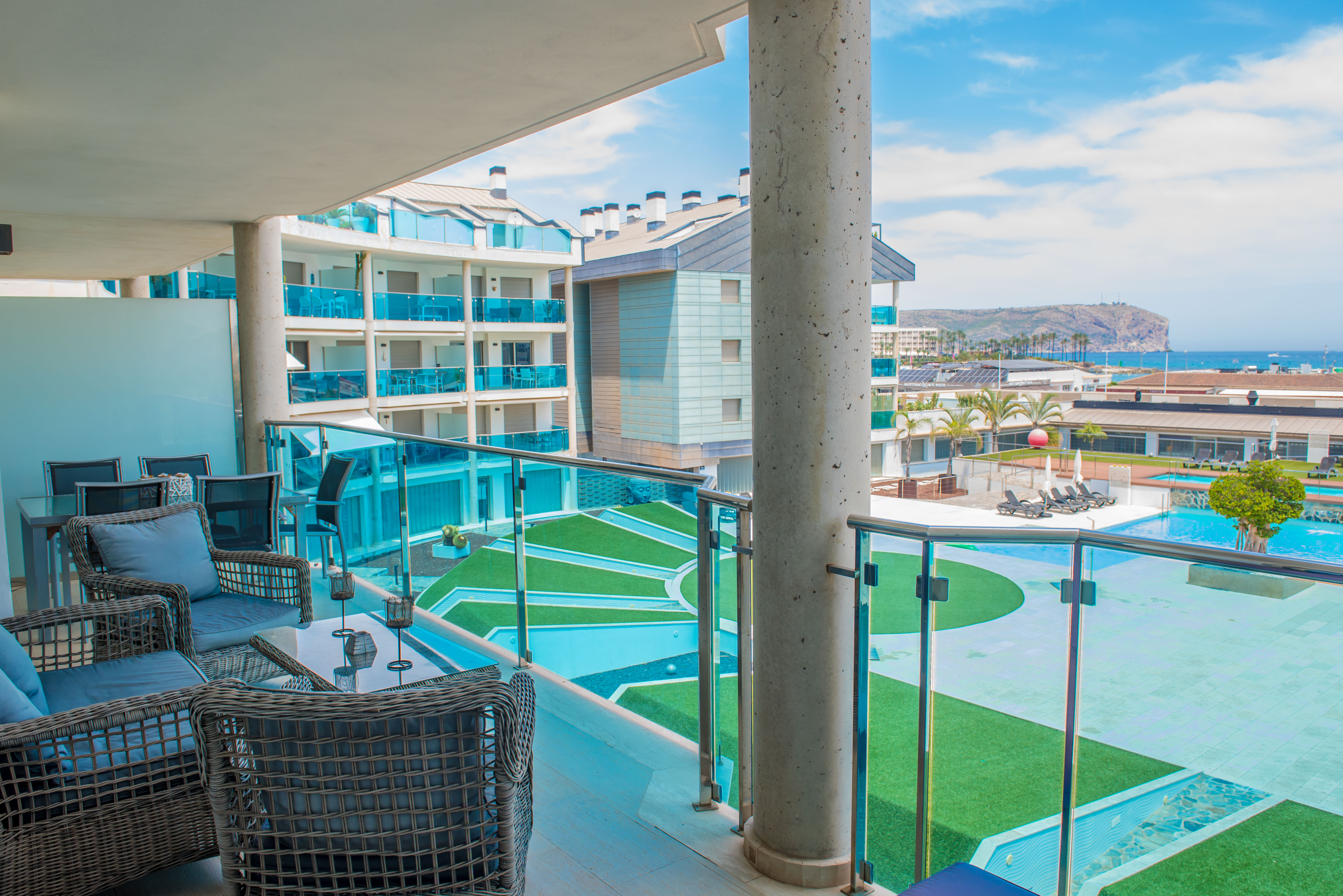 Vaste terrasse privée aménagée avec mobilier d'extérieur en rotin, offrant une vue directe sur les piscines communes et la mer Méditerranée. Garde-corps en verre.