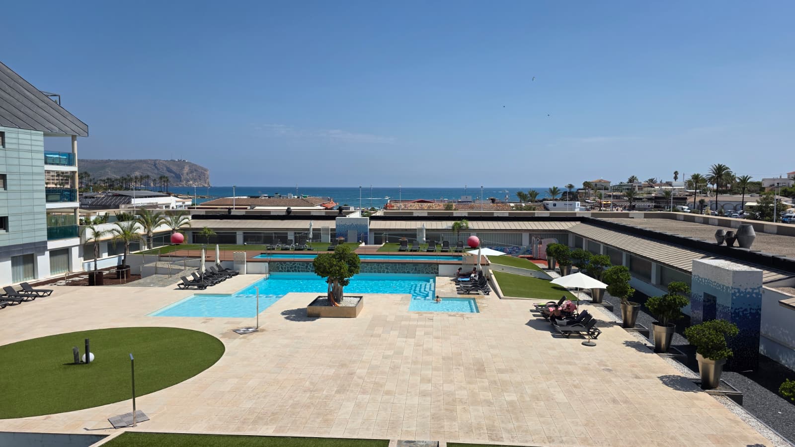 Vue aérienne de la vaste zone de piscines communes avec terrasses pavées, gazon artificiel et transats, en front de mer avec falaise lointaine.