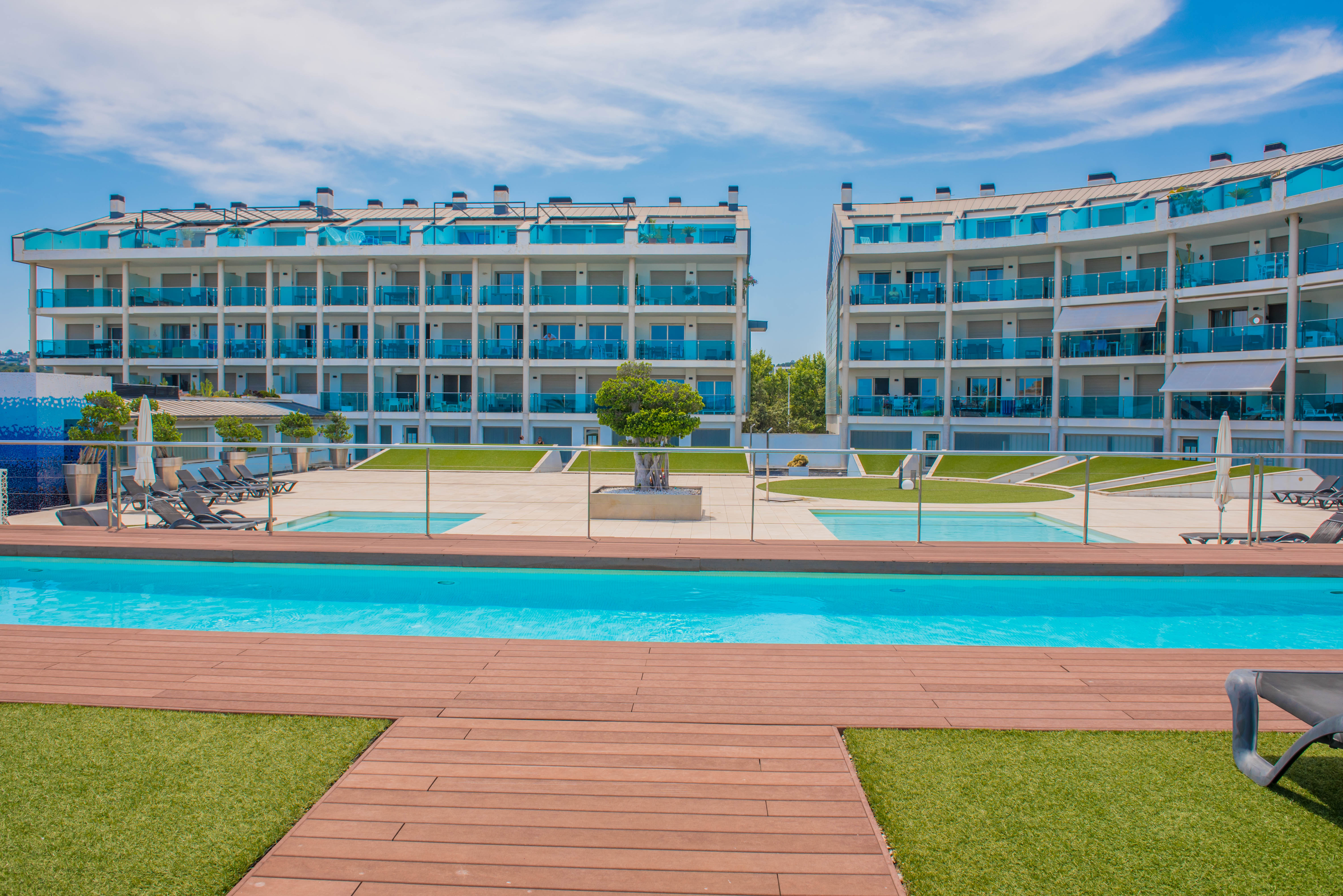 Vue de la piscine commune avec terrasse en bois et pelouse artificielle, encadrée par des bâtiments résidentiels modernes aux balcons en verre bleu.