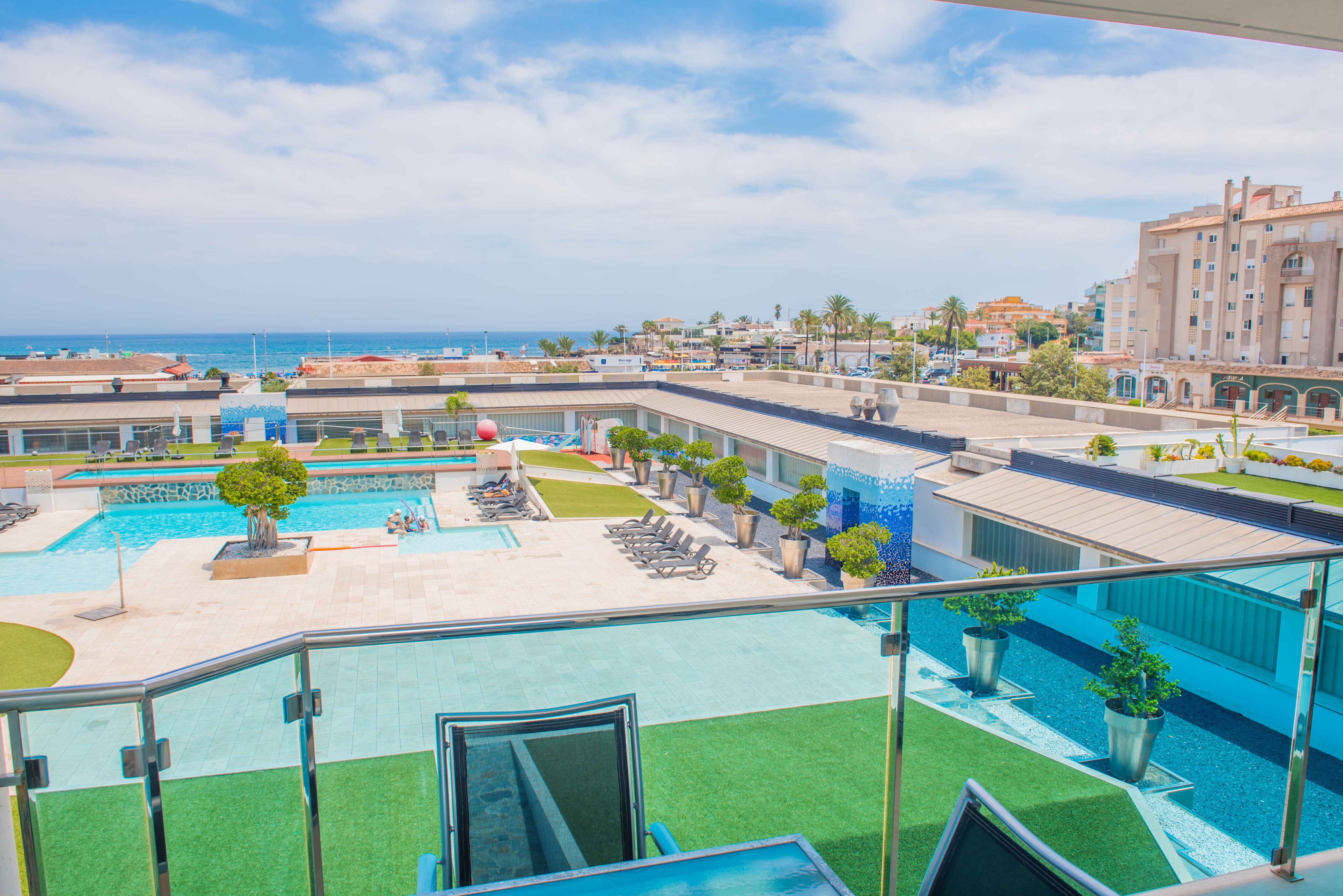 Vue en hauteur depuis la terrasse avec balustrade en verre, présentant la piscine commune, les zones de gazon artificiel et la mer Méditerranée au loin.