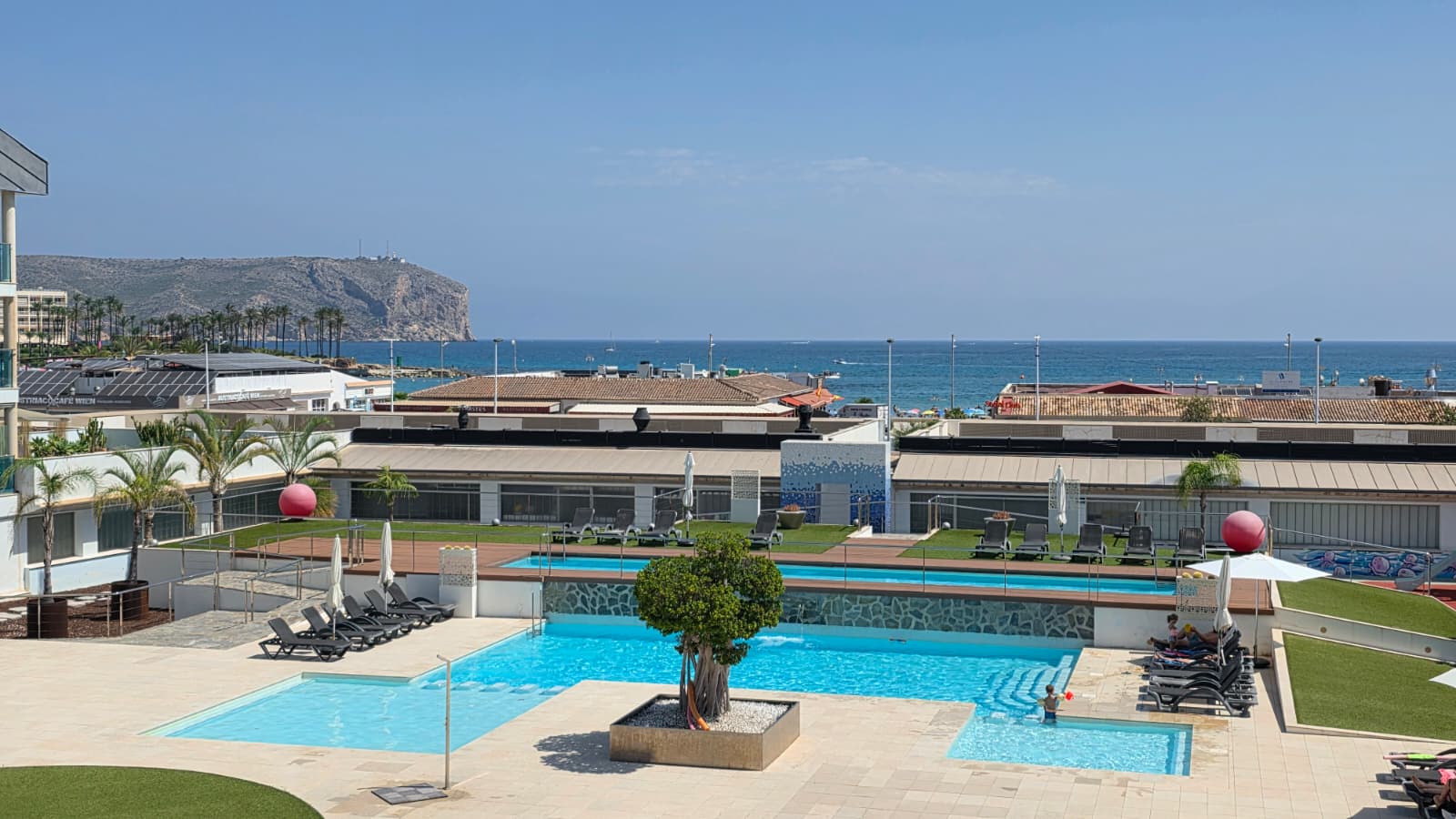 Vue panoramique de l'espace piscine à plusieurs niveaux avec un arbre central, entouré de chaises longues et de gazon artificiel, surplombant la mer Méditerranée et des falaises lointaines.