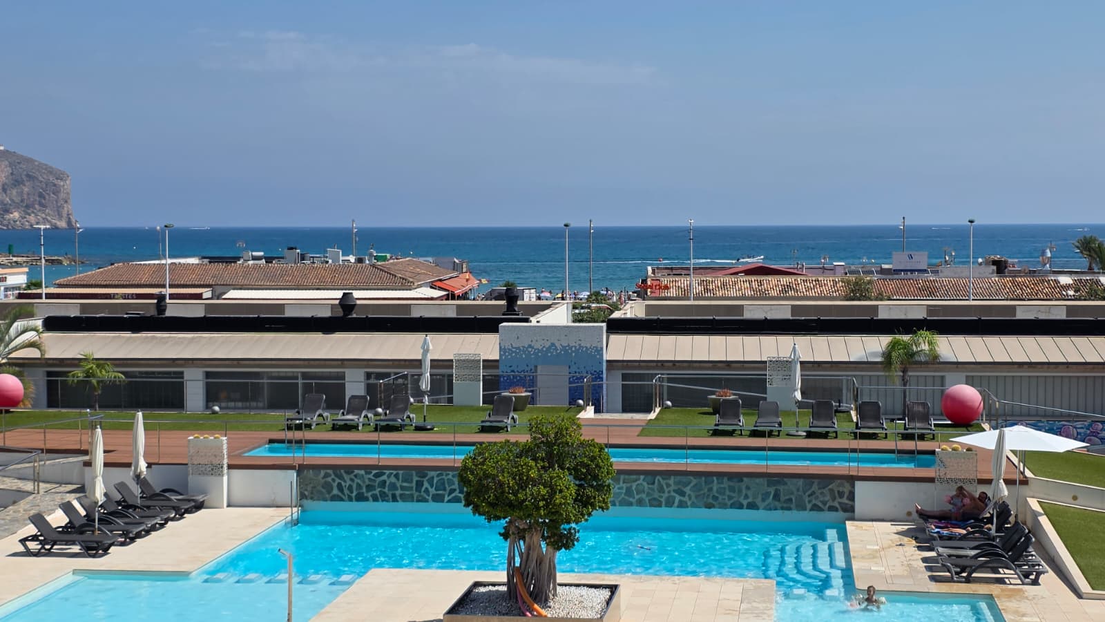 Vue panoramique sur les piscines à plusieurs niveaux du complexe, avec transats, gazon artificiel et vues directes sur la mer Méditerranée et la côte lointaine.
