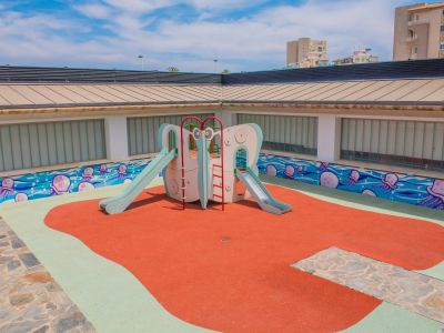 Aire de jeux pour enfants lumineuse avec structure de toboggans sur un sol en caoutchouc rouge et vert. Un mur décoré d'une fresque de méduses anime l'espace, sous des panneaux de verre dépoli.