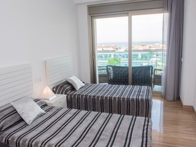 Chambre lumineuse avec deux lits simples, literie rayée et table de chevet. Des portes coulissantes mènent à un balcon avec sièges extérieurs et vue partielle sur la mer. Sol poli.