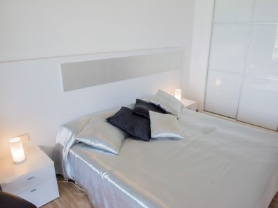 Chambre moderne avec lit aux draps satinés argentés, tête de lit blanche et tables de chevet assorties avec lampes cylindriques. Armoire coulissante en verre dépoli.