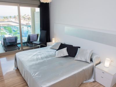 Chambre principale moderne avec grand lit, literie satinée argentée, carrelage brillant et portes coulissantes vers la terrasse offrant une vue sur la mer et la piscine.