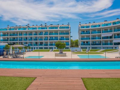 Complexe résidentiel moderne composé de deux bâtiments avec balcons incurvés en verre bleu, entourant une grande piscine commune avec terrasse en bois élégante et gazon artificiel.