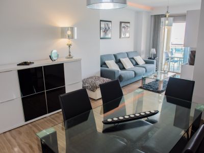 Espace de vie et salle à manger ouvert avec table en verre, chaises noires et canapé bleu. Le sol carrelé et un buffet blanc moderne complètent l'ensemble, menant à une terrasse.