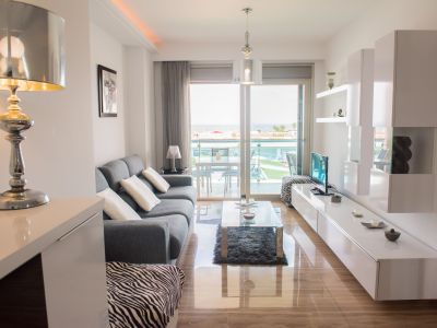 Salon moderne avec sols en marbre poli, s'ouvrant sur une terrasse offrant des vues sur la piscine commune et la mer. Canapé gris et meubles blancs contemporains.