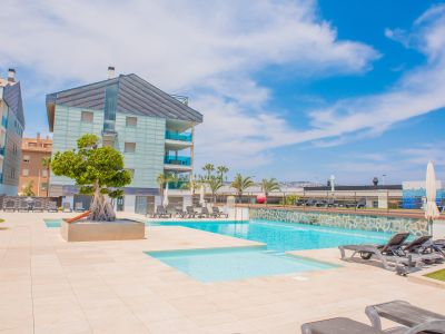 Vaste espace piscine commun avec deux bassins distincts, pavage clair et nombreuses chaises longues. Des bâtiments résidentiels modernes aux balcons vitrés bleus surplombent cette zone ensoleillée.