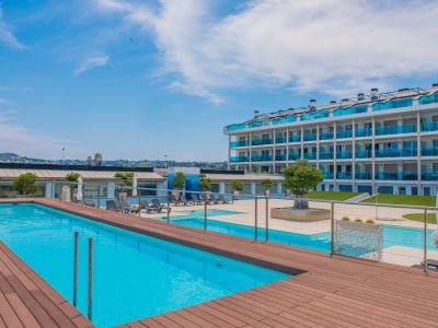 Vaste espace piscine commun avec une piscine d'un bleu éclatant bordée d'une terrasse en bois chaleureuse. L'immeuble moderne aux balcons en verre bleu distinctifs surplombe cet espace extérieur serein.
