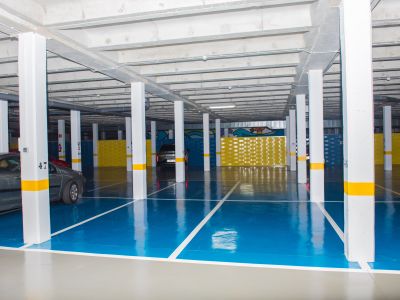 Vaste garage souterrain avec sol en résine bleu et gris, piliers blancs à bandes jaunes et plafond en béton apparent. Des places de stationnement et des panneaux décoratifs jaunes sont visibles.