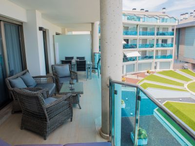 Vaste terrasse couverte avec salon en rotin, table basse en verre et carrelage clair. Vue sur les jardins communs modernes et un bâtiment voisin aux balcons en verre bleu.