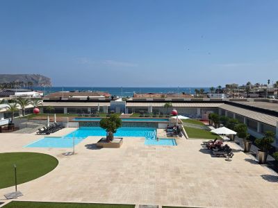 Vue aérienne de la vaste zone de piscines communes avec terrasses pavées, gazon artificiel et transats, en front de mer avec falaise lointaine.
