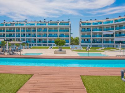 Vue de la piscine commune avec terrasse en bois et pelouse artificielle, encadrée par des bâtiments résidentiels modernes aux balcons en verre bleu.