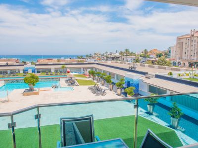 Vue en hauteur depuis la terrasse avec balustrade en verre, présentant la piscine commune, les zones de gazon artificiel et la mer Méditerranée au loin.