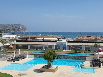 Vue panoramique de l'espace piscine à plusieurs niveaux avec un arbre central, entouré de chaises longues et de gazon artificiel, surplombant la mer Méditerranée et des falaises lointaines.